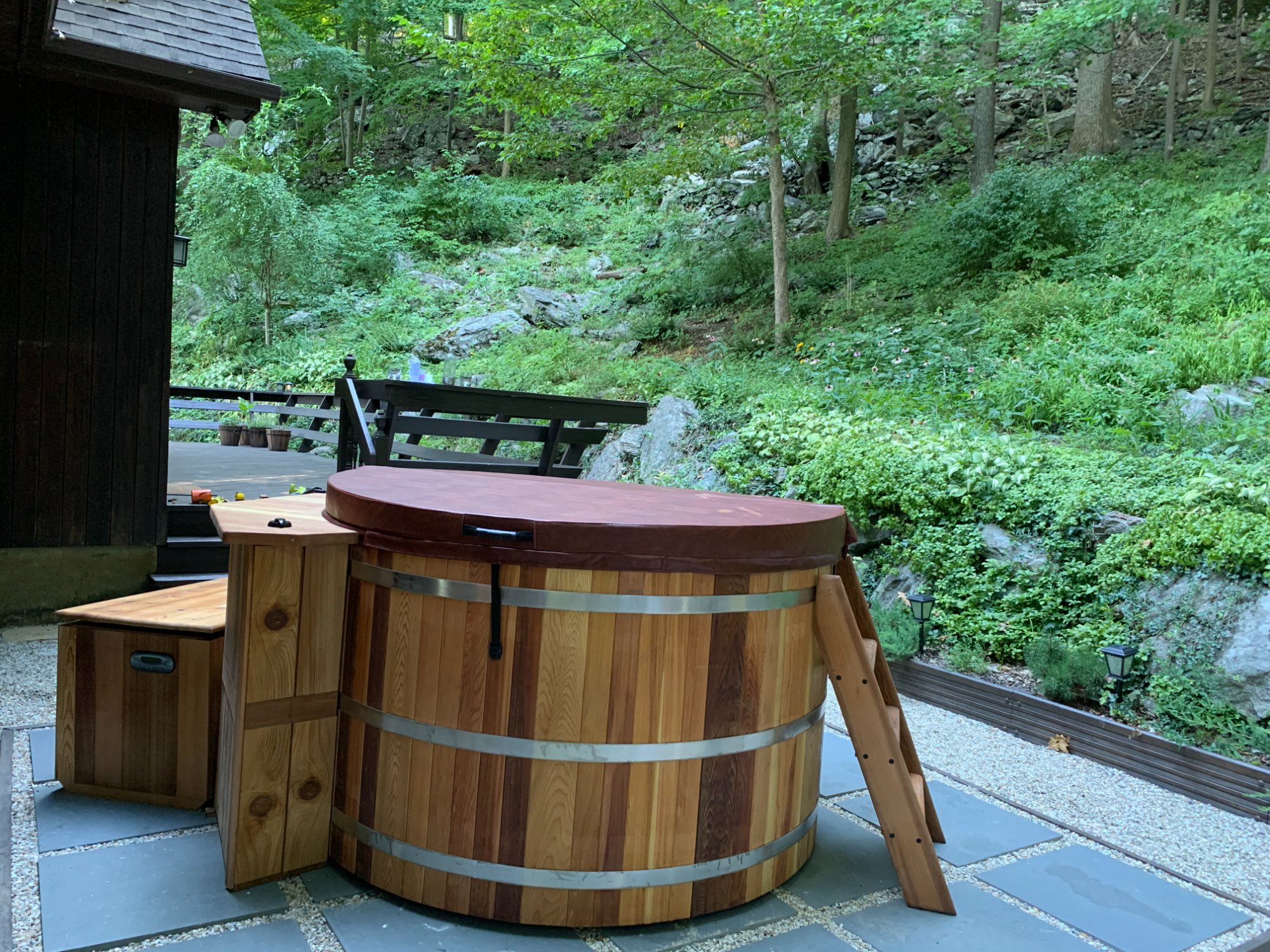 cedar hot tub with cover on a stone patio against a wooded hillside