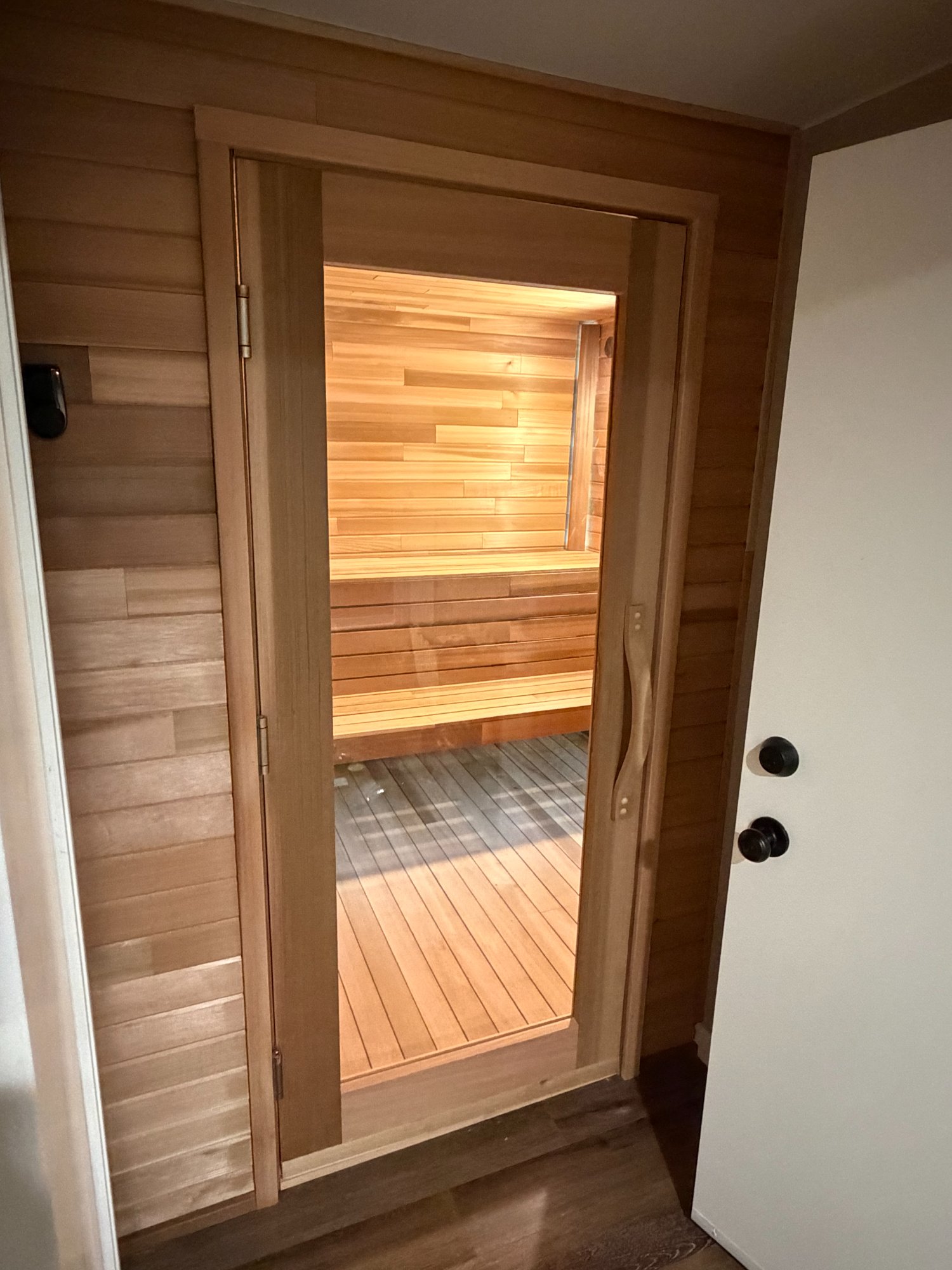 Custom cedar sauna glass door framed in cedar paneling, glowing warm interior visible from hallway