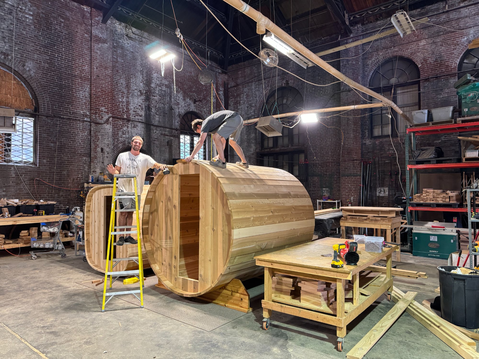 Two craftsmen building a large cedar barrel sauna in the Satori Cedar Spas workshop with arched brick windows